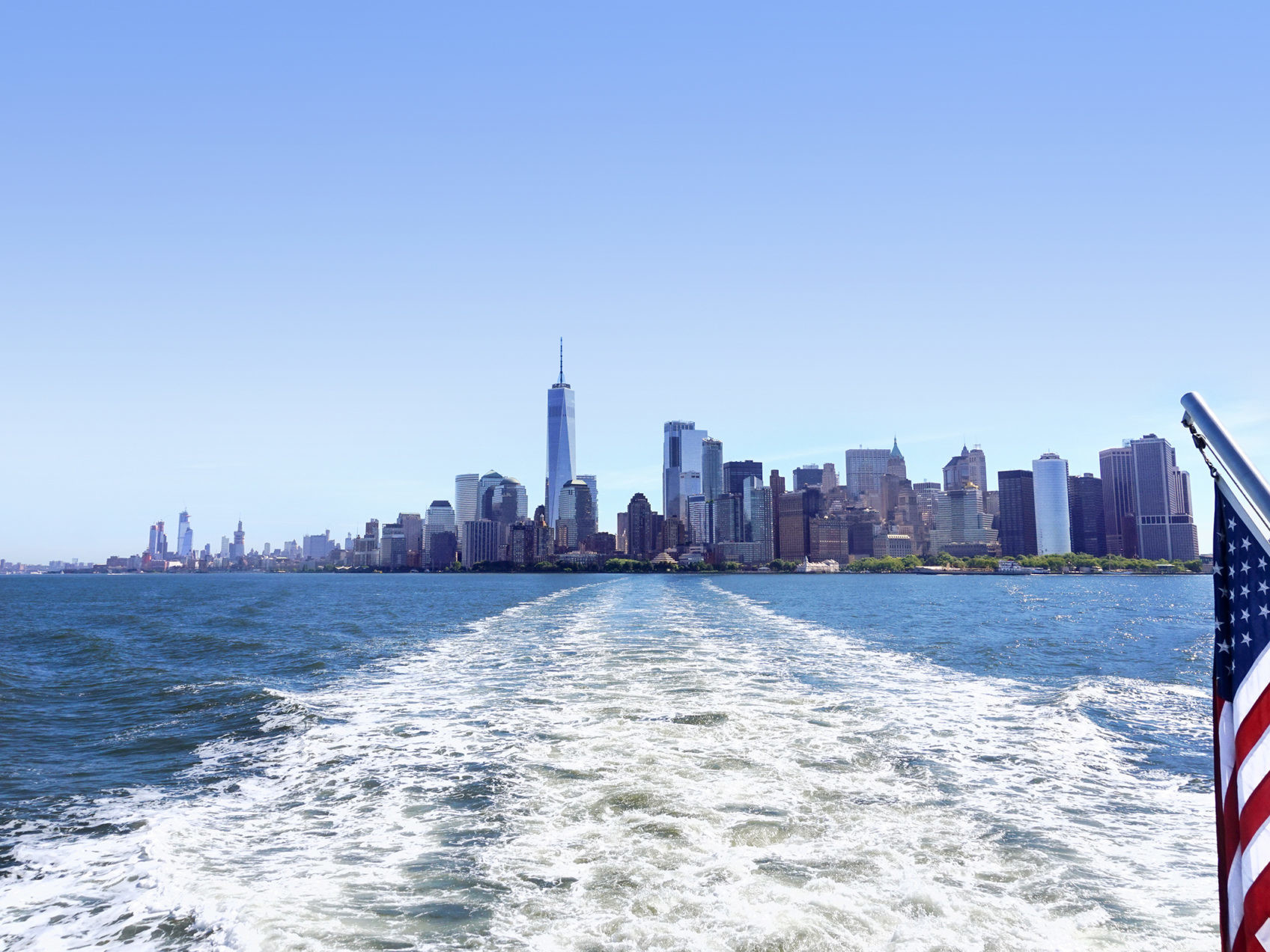 Lower Manhattan view from cruise ship or yacht with the flag of the United States of America in New York. Scenic view to NY Downtown and city centre. Staten Island Ferry. Tourists cruising in boat.