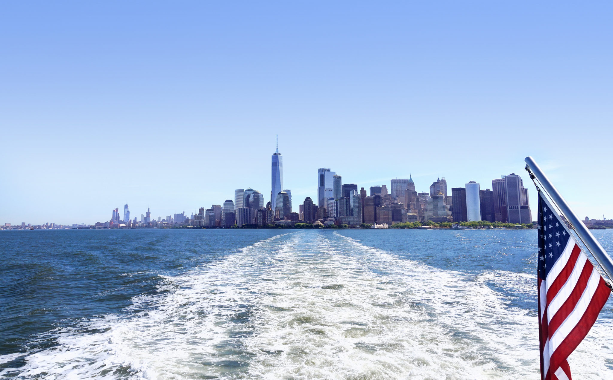 Lower Manhattan view from cruise ship or yacht with the flag of the United States of America in New York. Scenic view to NY Downtown and city centre. Staten Island Ferry. Tourists cruising in boat.